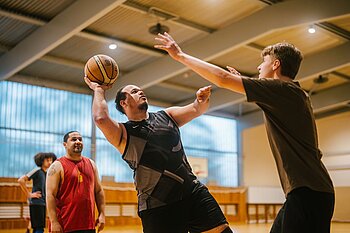 Zwei Männer spielen Basketball in einer Sporthalle, wobei einer den Ball hält und versucht, einen Wurf gegen die Verteidigung des anderen zu machen.