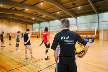 In einer Sporthalle beobachtet ein Trainer, der ein Volleyball in der Hand hält und ein T-Shirt mit der Aufschrift "Hochschule für Technik und Wirtschaft Berlin" trägt, eine Gruppe von Spielern beim Training.